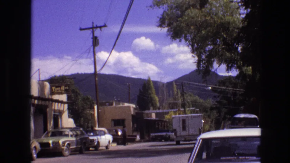 1973 New Mexico frame showing a white camper continuing along a small-town road past parked cars, storefronts, trees, utility wires, and distant mountains.