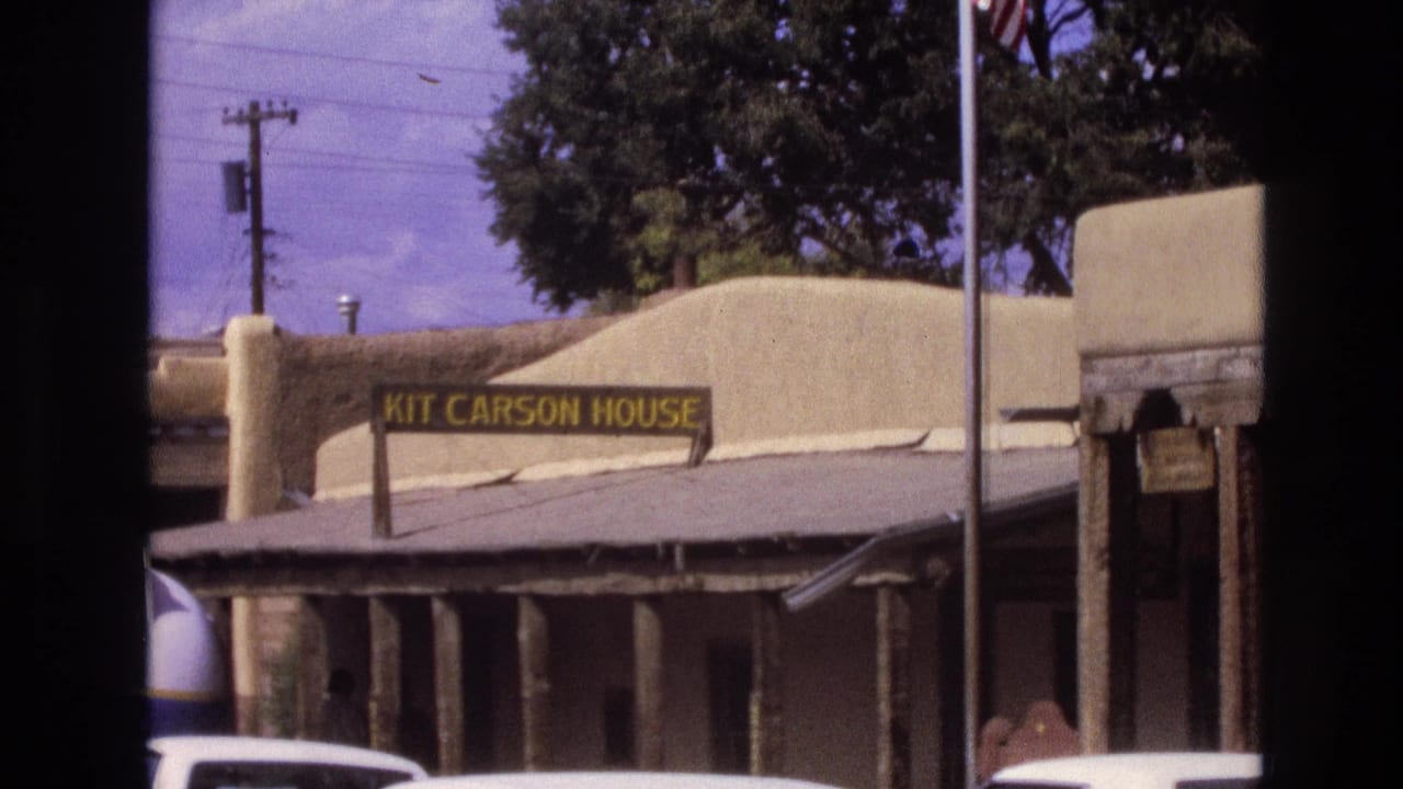1973 New Mexico home-movie frame showing the Kit Carson House sign on an adobe-style exterior with parked cars and trees nearby.