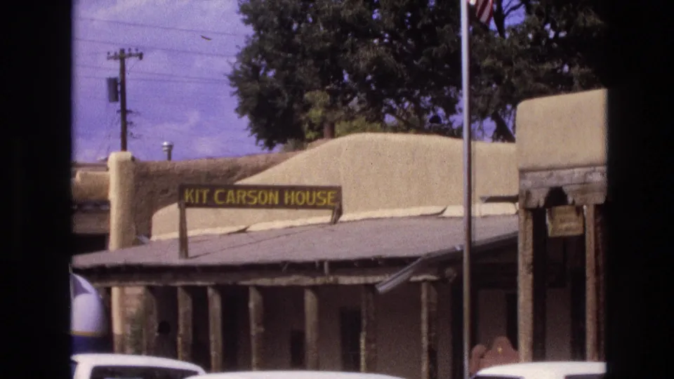 1973 New Mexico home-movie frame showing the Kit Carson House sign on an adobe-style exterior with parked cars and trees nearby.