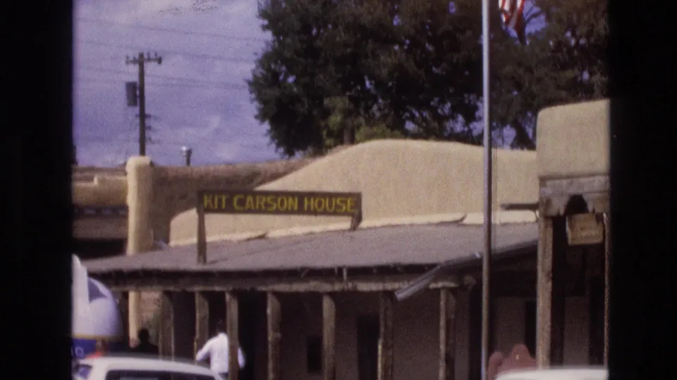 1973 New Mexico frame of the Kit Carson House exterior, with the yellow sign visible above the porch and a person walking near parked cars.
