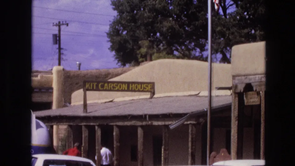 1973 New Mexico home-movie frame showing the Kit Carson House sign, adobe exterior, porch posts, parked car roofs, and pedestrians partly visible near the street.