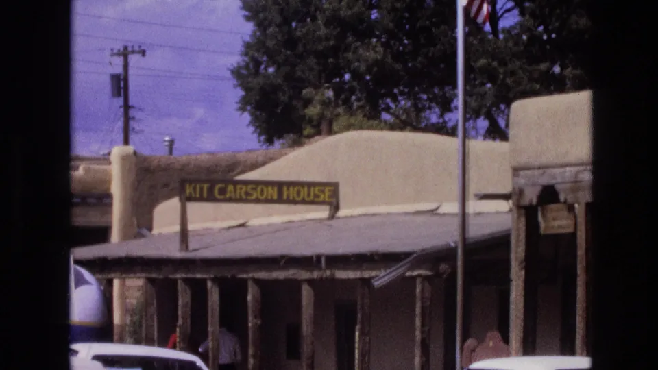 1973 New Mexico frame of the Kit Carson House exterior, with its sign, adobe-style wall, shaded porch, parked cars, and a person near the entrance area.