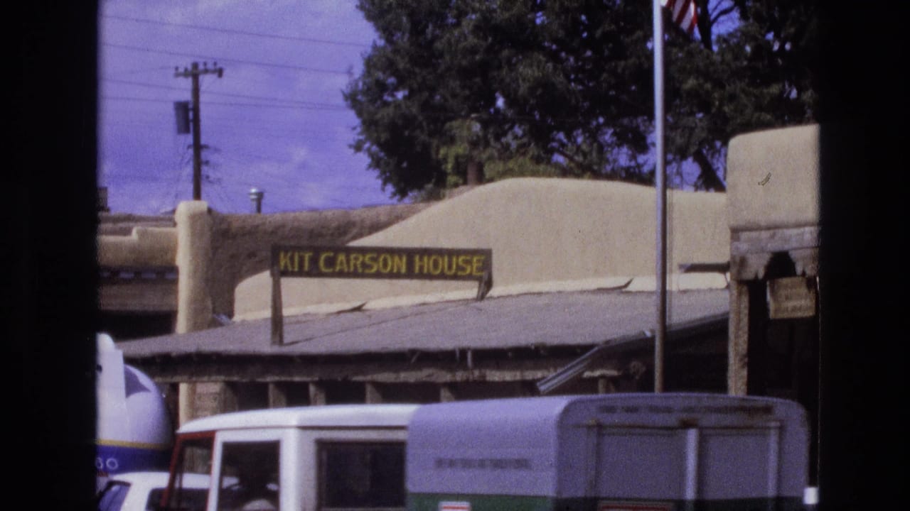 Adobe-style Kit Carson House exterior with sign, street vehicles, and a trailer in front.