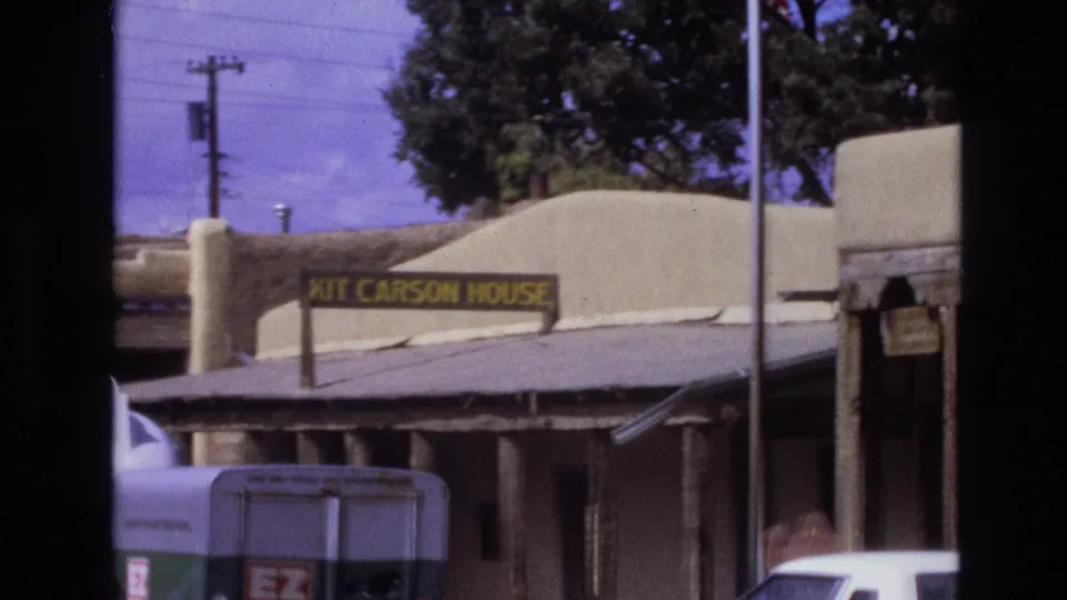 1973 New Mexico 8mm frame showing a closer view of the Kit Carson House sign and adobe exterior, partly obscured by a trailer and a white vehicle at street level.