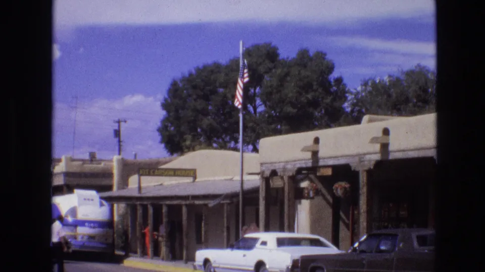 Wider 1973 New Mexico frame of the Kit Carson House streetscape, showing an American flag, adobe-style buildings, parked cars, trees, and small human figures near the entrance area.