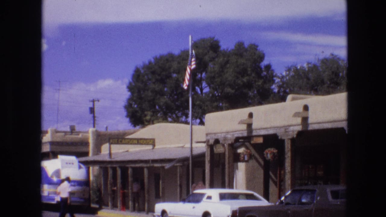 1973 New Mexico street view with adobe-style storefronts, a Kit Carson House sign, an American flag, classic cars, a tour bus, and pedestrians on the sidewalk.