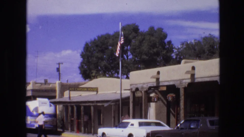1973 New Mexico street view with adobe-style storefronts, a Kit Carson House sign, an American flag, classic cars, a tour bus, and pedestrians on the sidewalk.