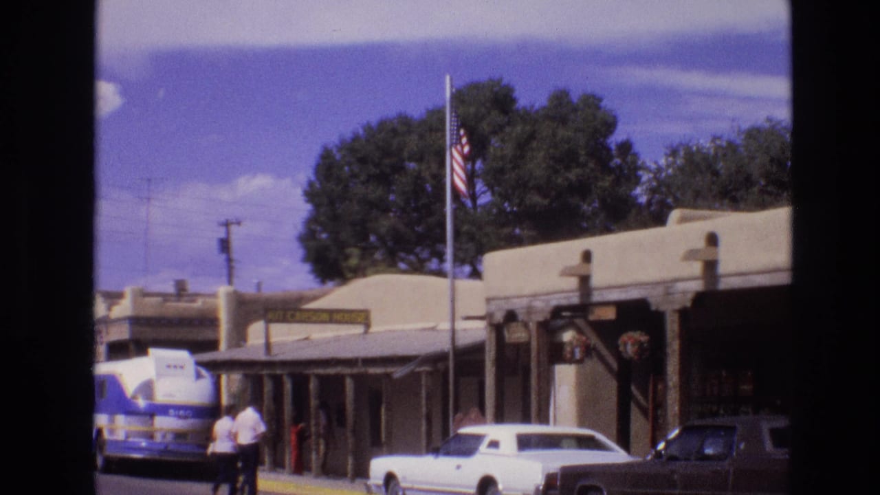 Adobe-style town buildings with a flagpole, parked cars, a bus, and people walking near the curb.