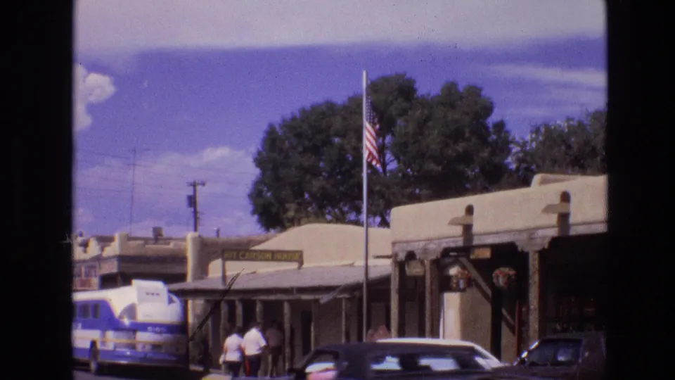 1973 New Mexico home-movie frame with a tour bus at left, Kit Carson House signage, adobe-style buildings, a flagpole, pedestrians, and period cars along the street.