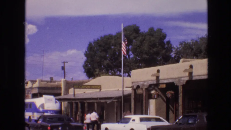 1973 New Mexico street scene with an American flag above adobe-style buildings, a blue-and-white tour bus, two pedestrians, a white classic car, and a dark vehicle near the curb.