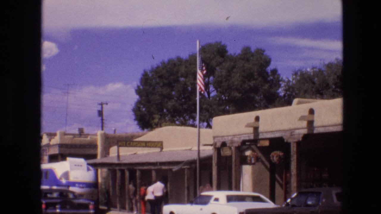 1973 New Mexico street view with an American flag on a pole outside adobe-style buildings, a blue-and-white bus, classic cars, and people on the sidewalk.