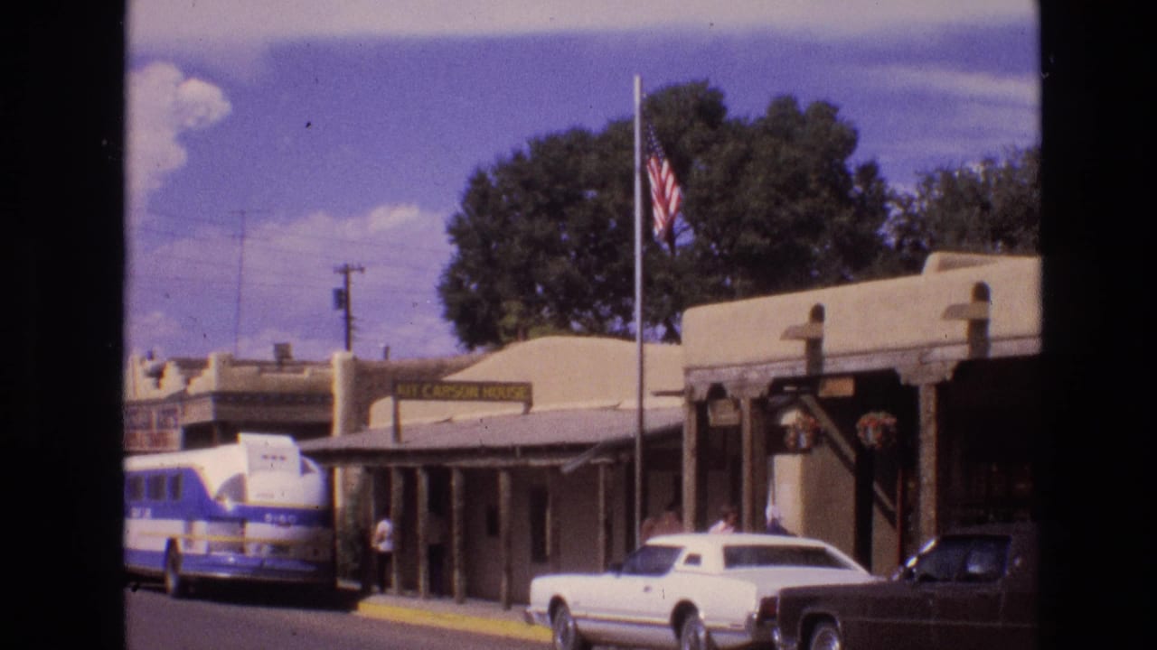 Flag outside a low adobe-style building with a bus, white car, dark pickup, and pedestrians nearby.