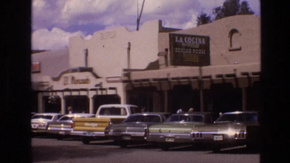 1973 New Mexico street scene showing adobe-style commercial facades, visible La Cocina and El Mercado signage, and several period cars parked along the curb.