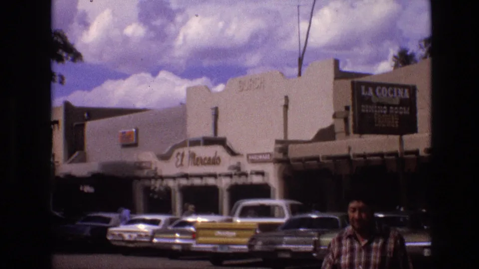 1973 New Mexico street view with La Cocina and El Mercado signage, rows of classic cars, a white pickup, adobe-style facades, and a man partly visible in the foreground.