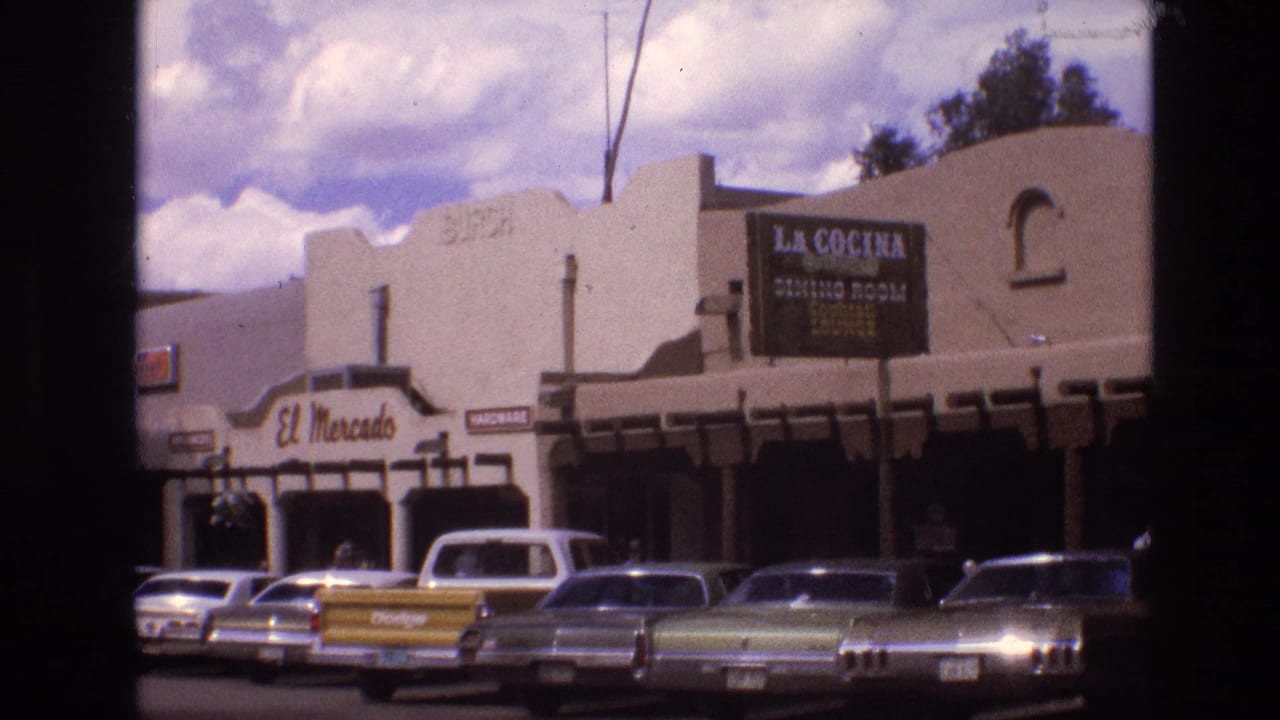 1973 New Mexico 8mm frame showing stucco commercial buildings with El Mercado and La Cocina signage, a yellow Dodge pickup, and several parked cars under a cloudy sky.