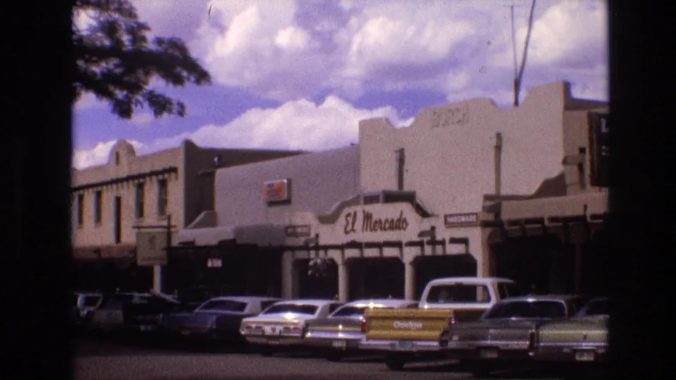 1973 New Mexico 8mm frame showing a wider angle of the stucco commercial block, the El Mercado sign, shaded storefronts, parked sedans, and the yellow Dodge pickup at the curb.