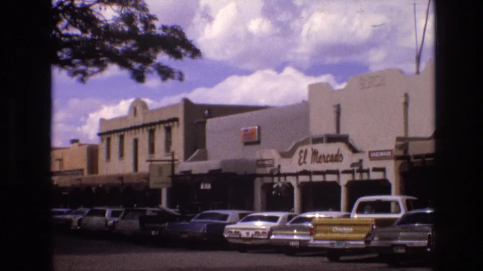 1973 New Mexico 8mm frame with the El Mercado storefront centered, classic cars parked in front, a yellow Dodge pickup at right, tree branches overhead, and bright clouds behind the building.
