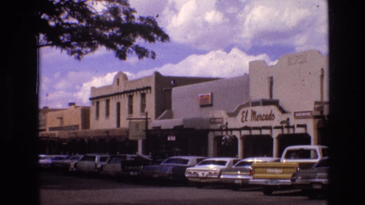 1973 New Mexico downtown street view with classic cars parked in front of adobe-style storefronts, including a visible El Mercado sign, framed by tree shade.