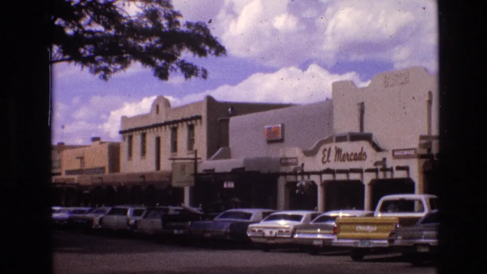 1973 New Mexico downtown street view with classic cars parked in front of adobe-style storefronts, including a visible El Mercado sign, framed by tree shade.