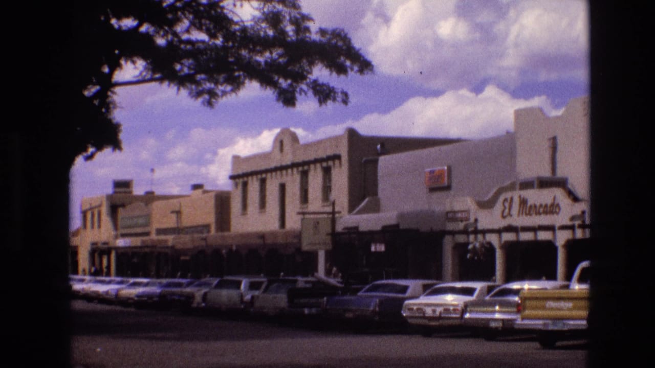 1973 New Mexico street scene showing parked American cars, adobe-style commercial buildings, El Mercado storefront signage, and dark tree branches across the upper frame.