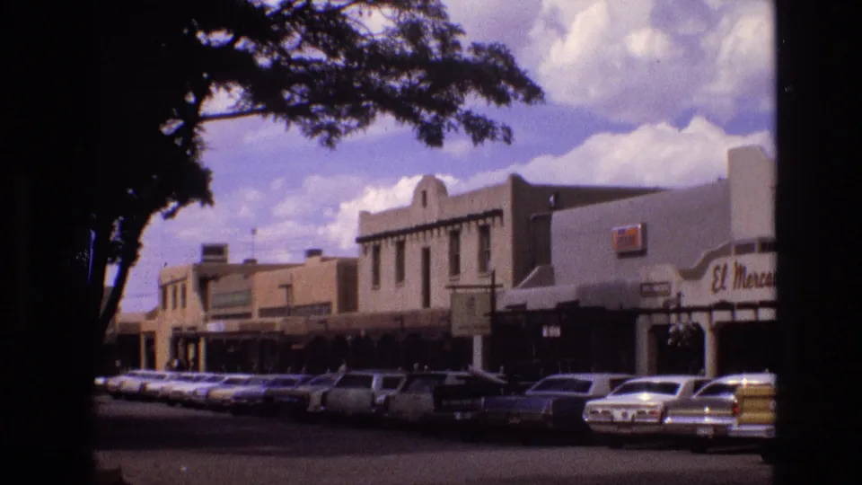 1973 New Mexico home-movie frame of a downtown commercial block with classic cars parked at the curb, shaded foreground trees, and a partial El Mercado storefront sign.