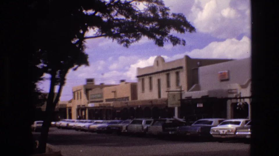 1973 New Mexico downtown street frame with a row of parked cars, adobe-style storefronts, tree trunks in the foreground, and bright clouds above.