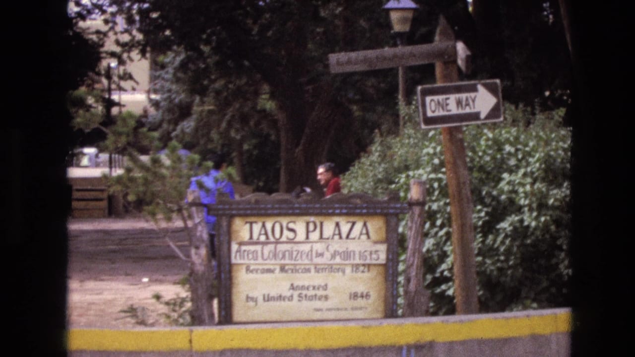 1973 New Mexico home-movie frame showing the Taos Plaza historical marker, a one-way sign, shrubs, trees, and distant pedestrians near a shaded path.