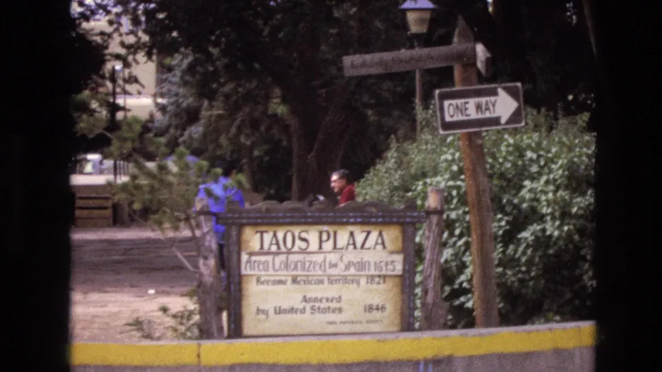 1973 New Mexico home-movie frame showing the Taos Plaza historical marker, a one-way sign, shrubs, trees, and distant pedestrians near a shaded path.
