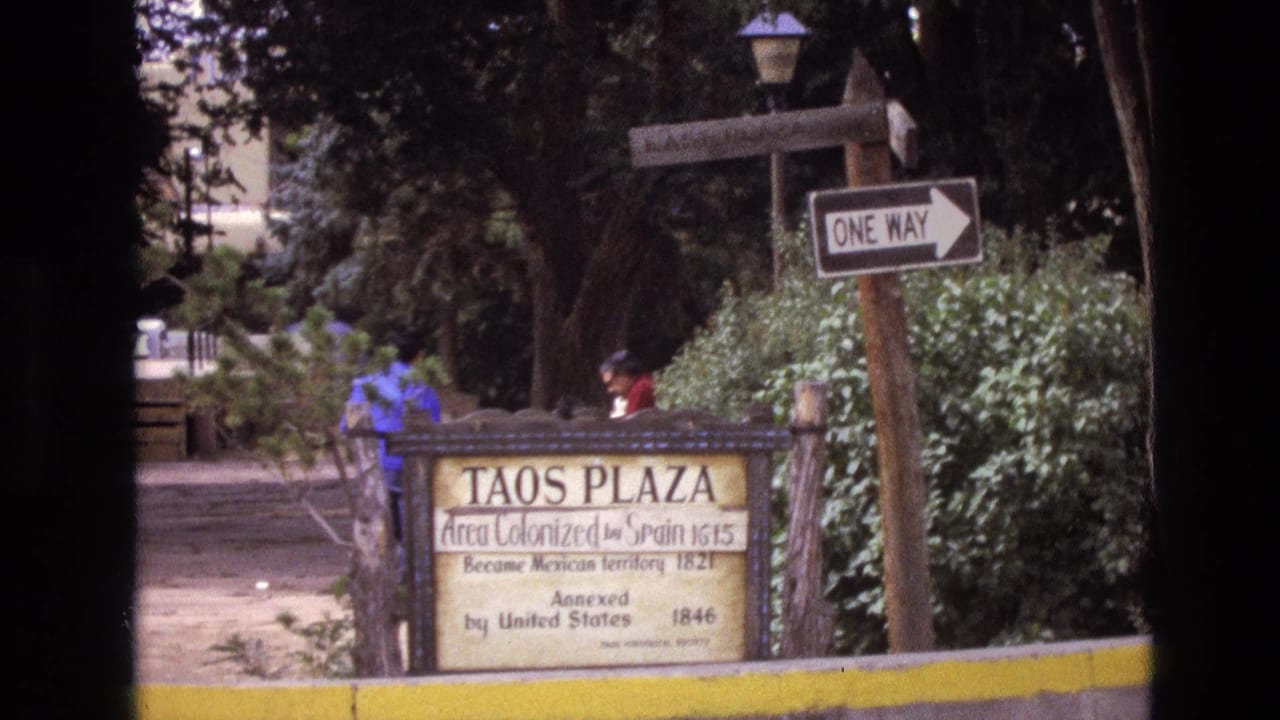 1973 New Mexico frame of the Taos Plaza marker partly framed by foliage, with a one-way street sign and distant people along the path.