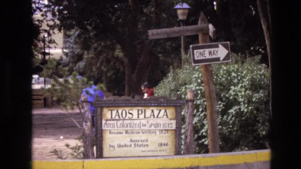 1973 New Mexico frame of the Taos Plaza marker partly framed by foliage, with a one-way street sign and distant people along the path.