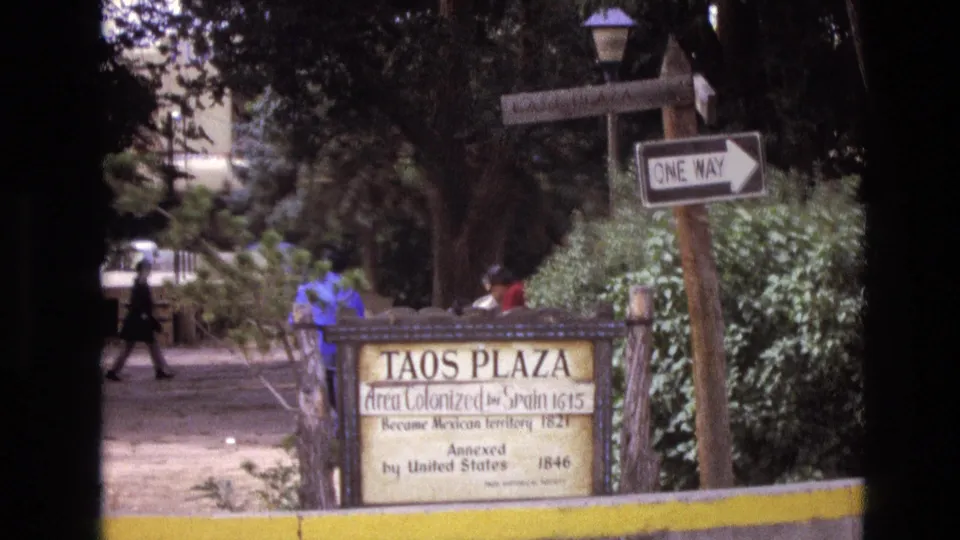 1973 New Mexico 8mm frame showing the Taos Plaza marker, yellow curb, shrubs, trees, and a pedestrian moving through the shaded plaza background.