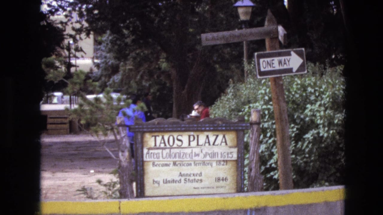 1973 New Mexico home-movie frame showing the Taos Plaza historical sign near a one-way sign, trees, shrubs, a yellow curb, and two people partly visible behind the marker.