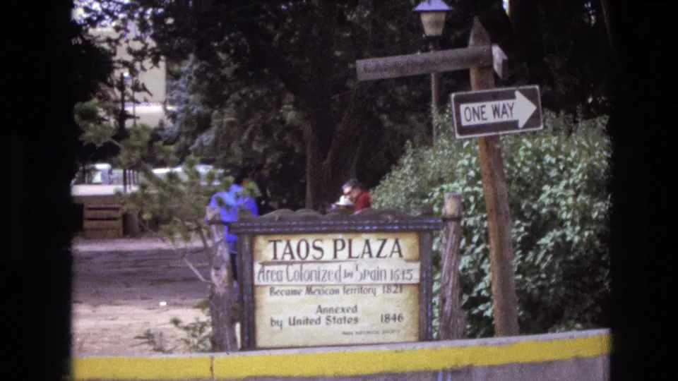 1973 New Mexico home-movie frame showing the Taos Plaza historical sign near a one-way sign, trees, shrubs, a yellow curb, and two people partly visible behind the marker.