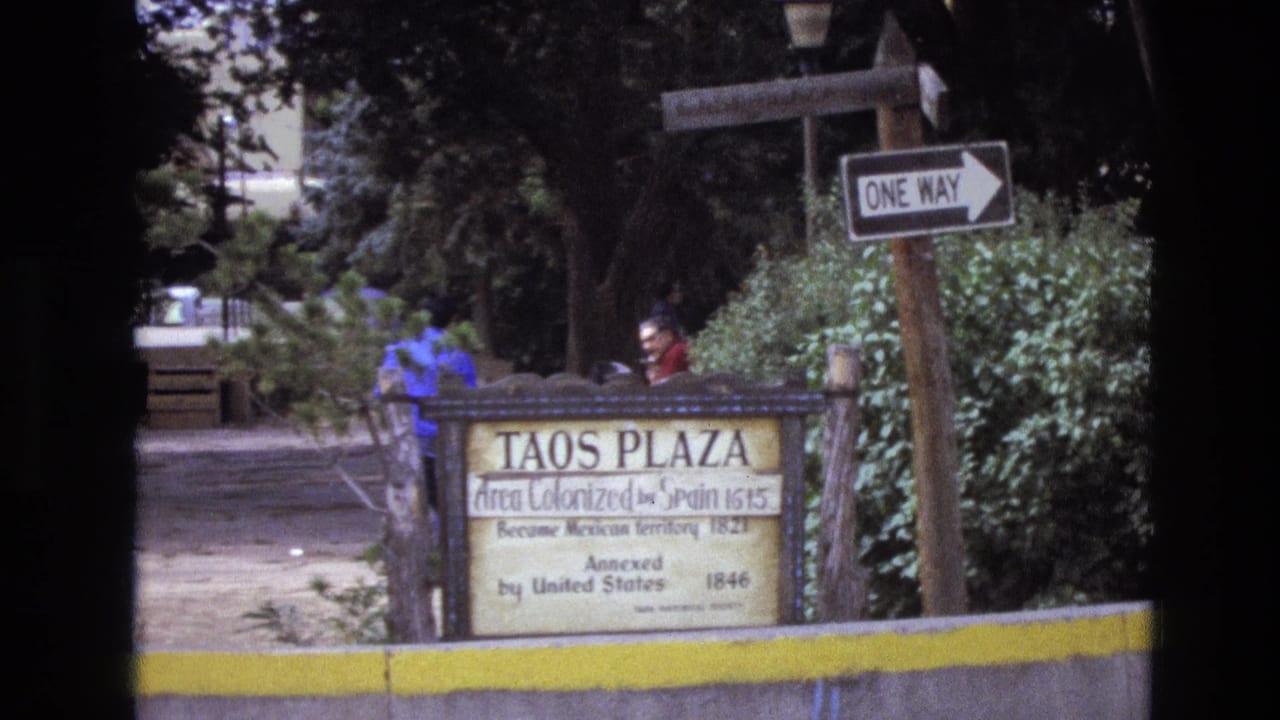 Taos Plaza sign framed by trees and shrubs with two people behind it.
