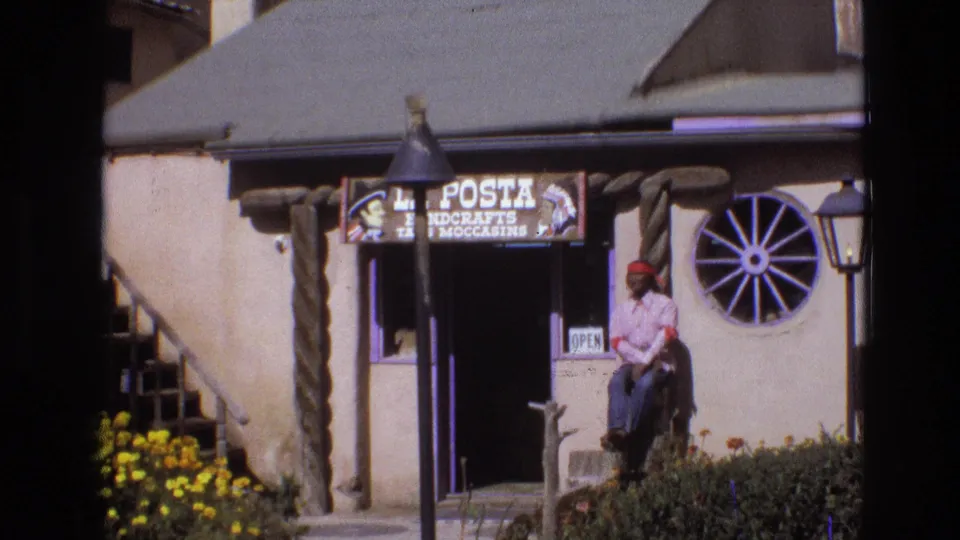 1973 New Mexico home-movie frame showing the La Posta Handcrafts storefront, a seated man in a red bandana and light shirt, an open sign, flowers, and a wagon-wheel wall decoration.