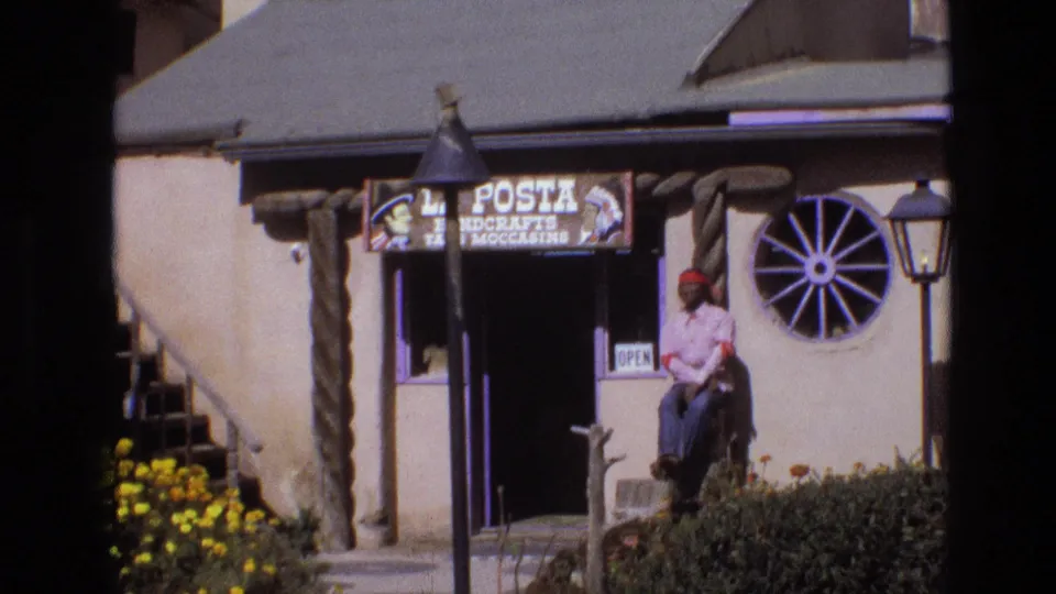 1973 New Mexico home-movie frame of the La Posta Handcrafts shopfront, with a seated man by the entrance, a wagon-wheel decoration, lamp posts, flowers, and an open sign.