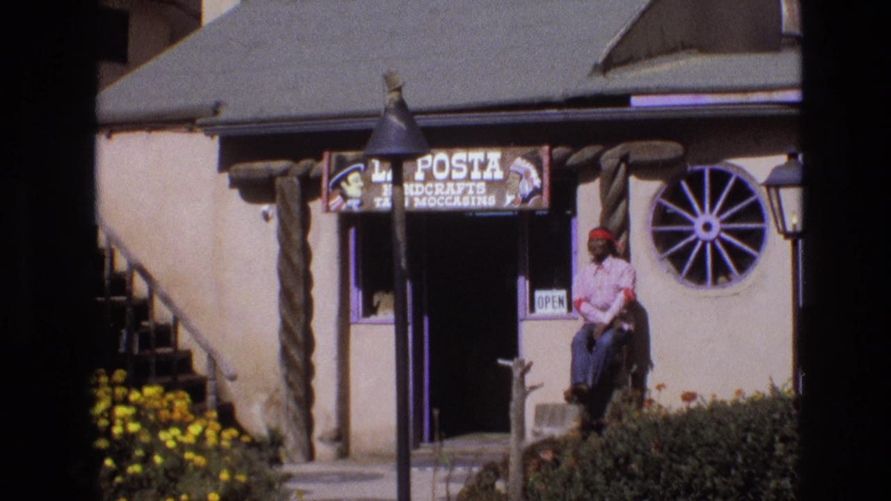 1973 New Mexico 8mm frame showing a Western-themed adobe-style shop exterior with a La Posta Handcrafts and Moccasins sign, wagon-wheel window, yellow flowers, and a seated man near the doorway.