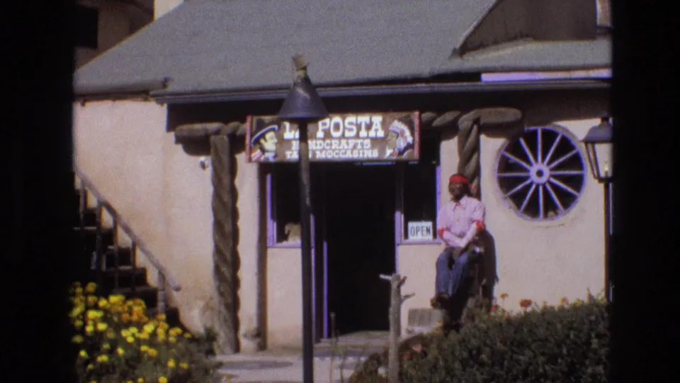 1973 New Mexico 8mm frame showing a Western-themed adobe-style shop exterior with a La Posta Handcrafts and Moccasins sign, wagon-wheel window, yellow flowers, and a seated man near the doorway.