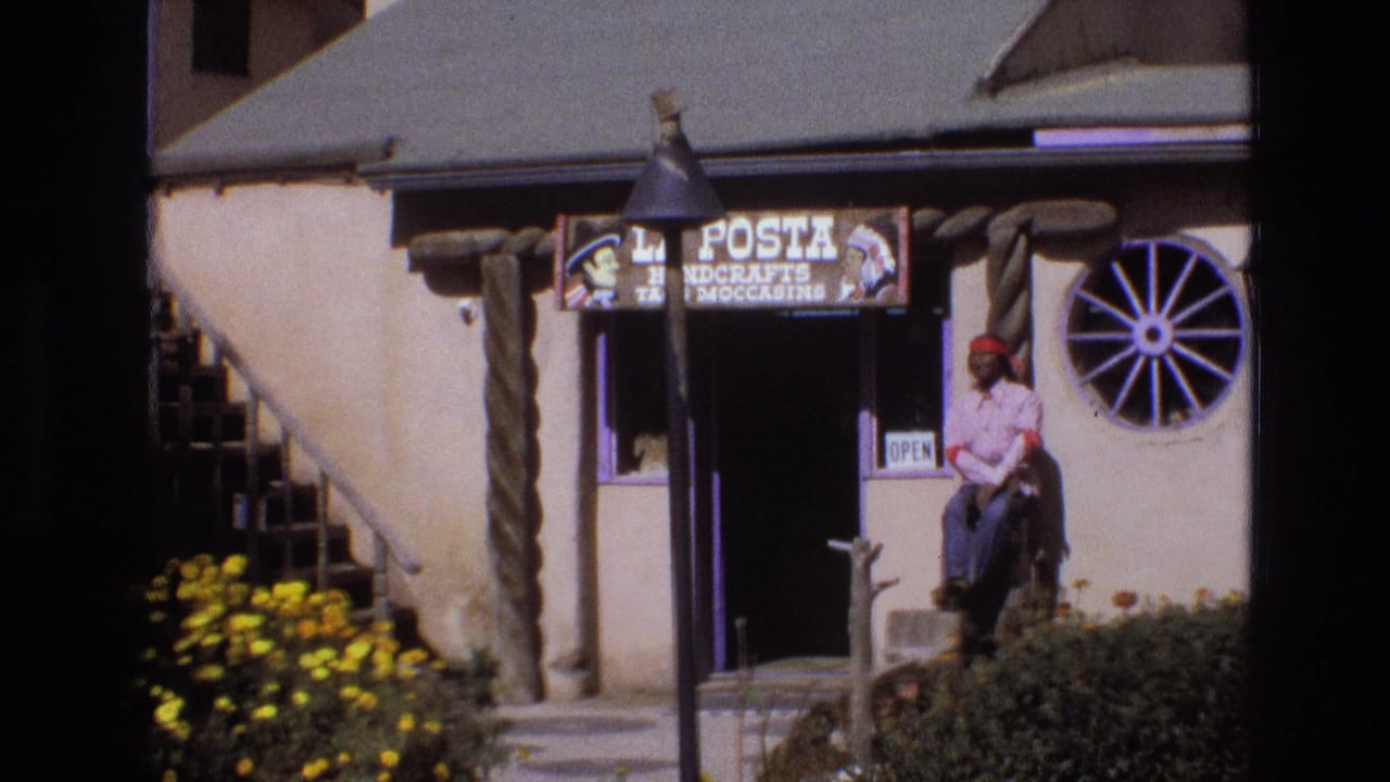 1973 New Mexico frame of the La Posta Handcrafts and Moccasins storefront, showing the open sign, round wagon-wheel window, rustic posts, and a seated man beside the entrance.