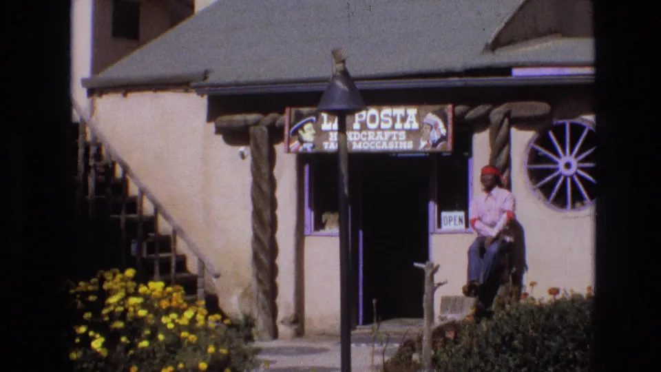 1973 New Mexico 8mm frame with a closer view of the La Posta sign, adobe-style entrance, wagon-wheel window, yellow flowers, and a seated man in a red bandana near the doorway.