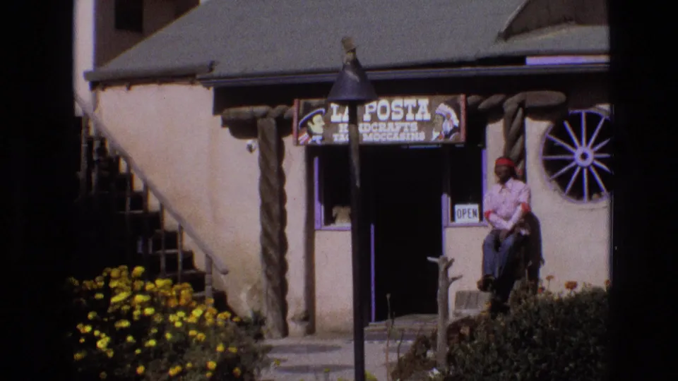 1973 New Mexico frame showing the La Posta Handcrafts and Moccasins exterior, including the wagon-wheel window, rustic adobe-style details, open doorway, foreground flowers, and seated man at the right edge.