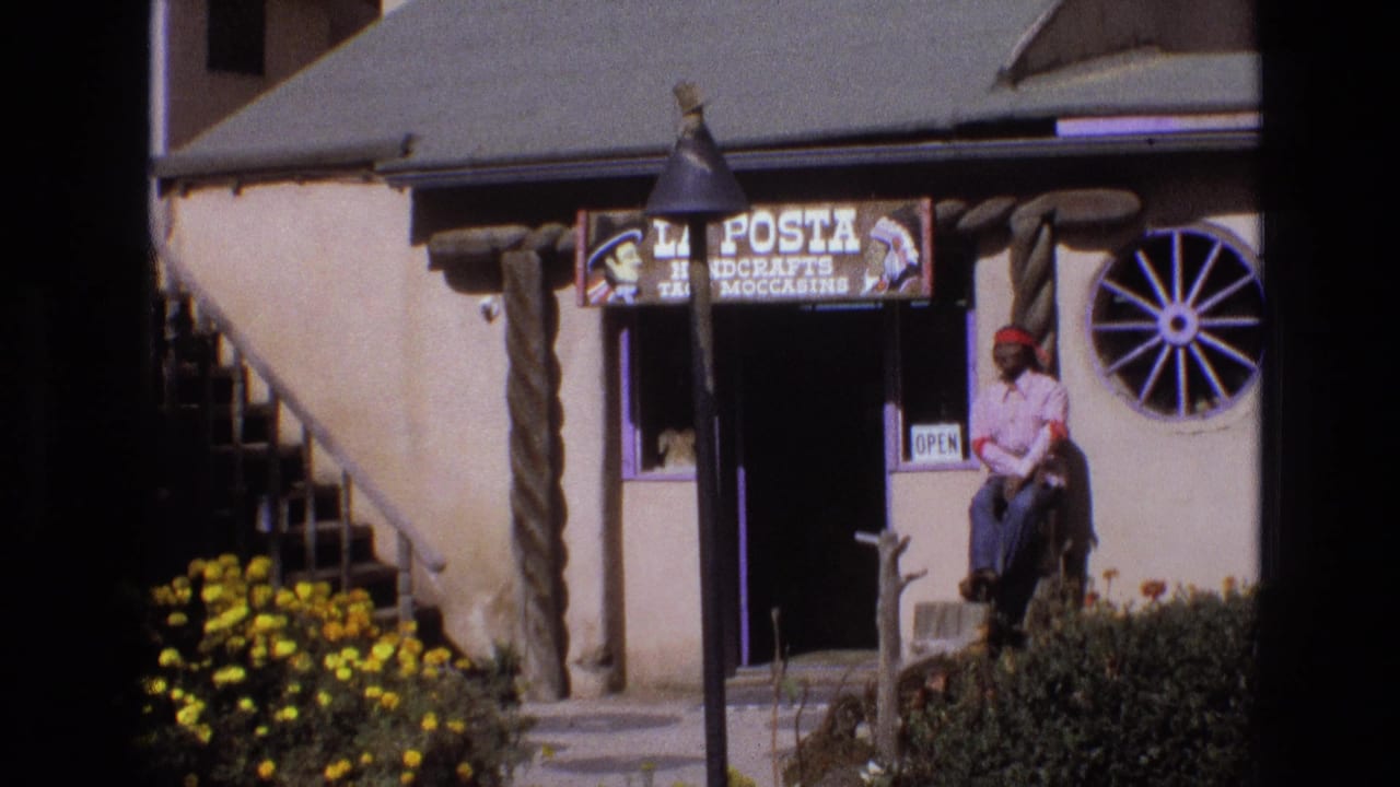 Adobe-style roadside shop with La Posta sign, open doorway, wagon-wheel window, flowers, and seated man.