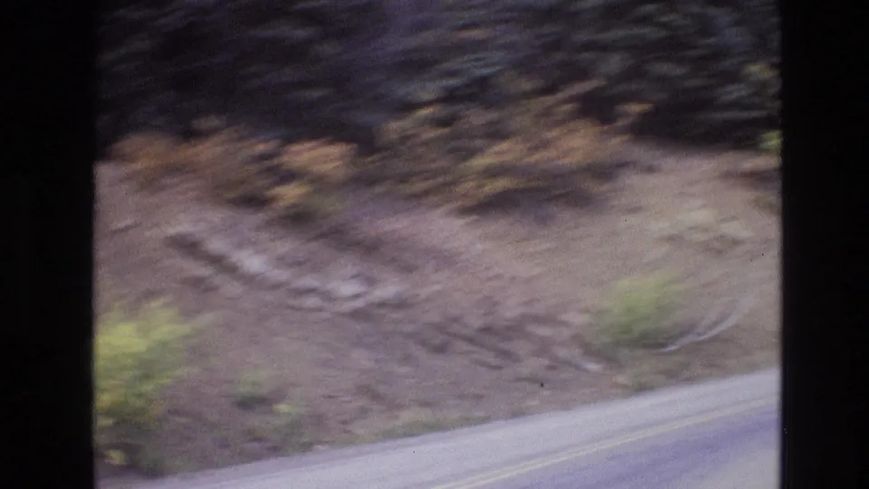 1973 New Mexico 8mm frame with a curving paved road, yellow center lines, rocky roadside slope, and evergreens passing in blur.