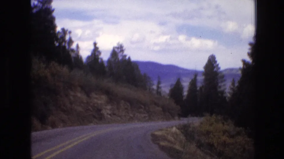 1973 New Mexico 8mm frame of a two-lane road curving through forested mountain terrain, with pine silhouettes, brush, distant hills, and soft film blur.