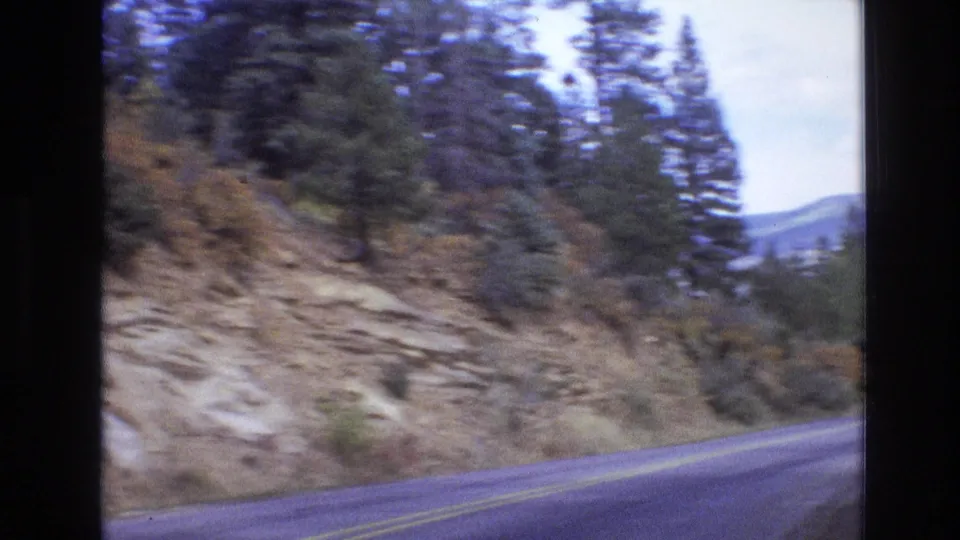 Rural New Mexico highway with double yellow lines curving beside a rocky, tree-lined slope, with distant hills visible at the right edge.