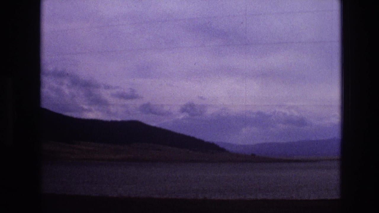 1973 New Mexico 8mm frame showing a silhouetted mountain ridge, distant hills, water, and an overcast purple-blue sky from a moving roadside view.