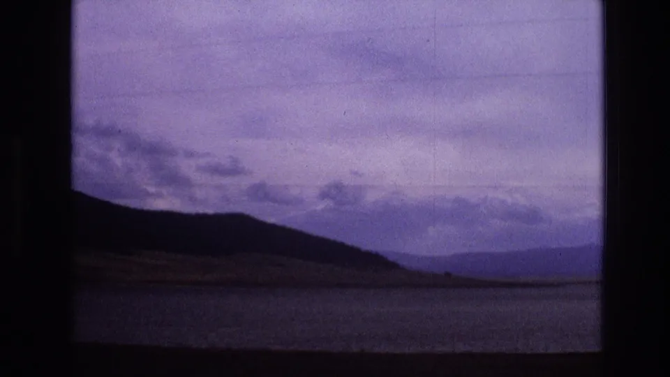 1973 New Mexico 8mm frame showing a silhouetted mountain ridge, distant hills, water, and an overcast purple-blue sky from a moving roadside view.