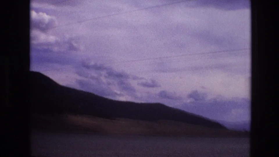 1973 New Mexico 8mm travel frame showing a dark hillside, water, distant low terrain, and layered overcast clouds with visible film grain.