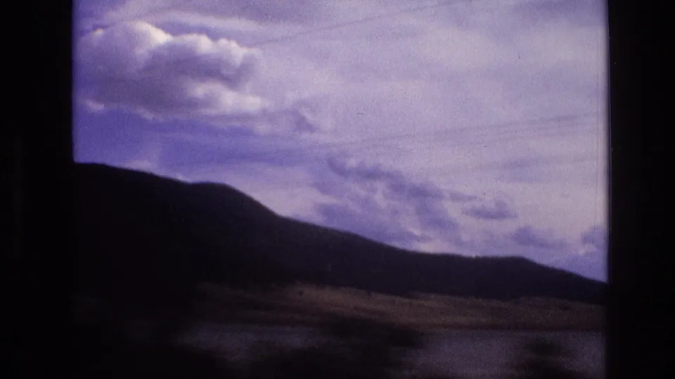 1973 New Mexico 8mm frame showing a silhouetted ridge, water at the base, bright cloud formations, and soft motion blur from a moving car.