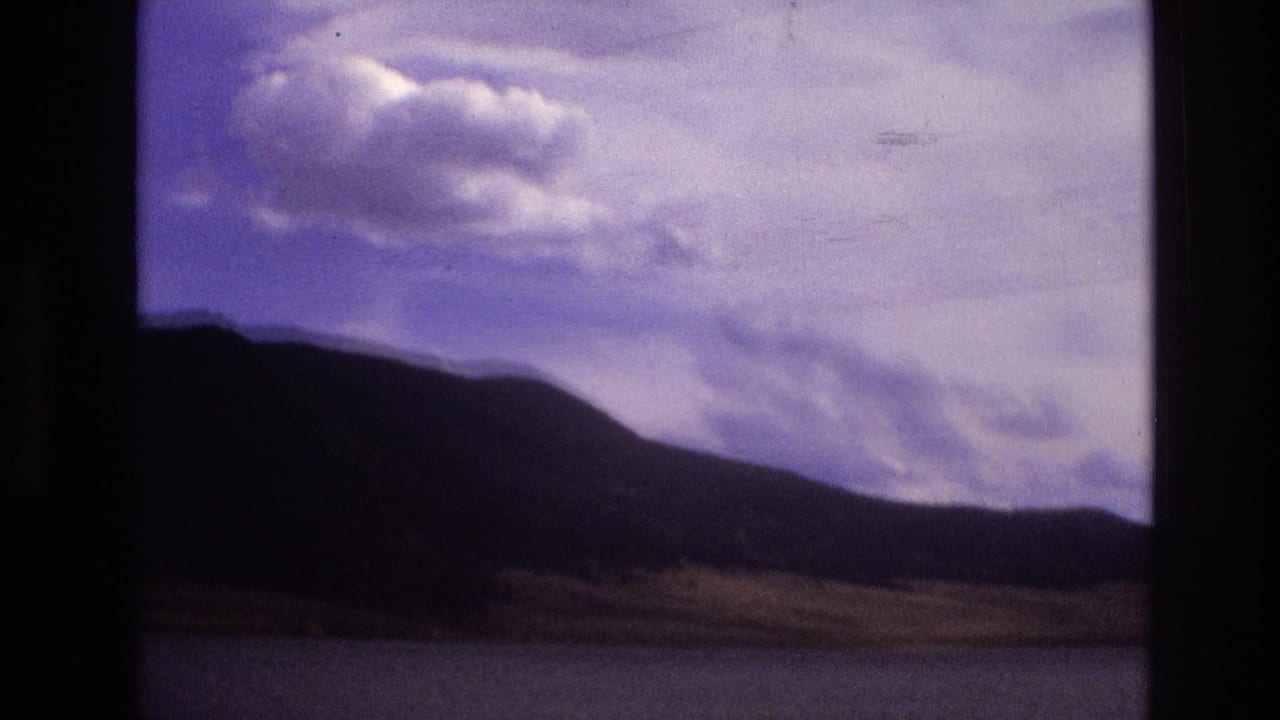 1973 New Mexico home-movie frame showing calm water in the foreground with dark mesa-like hills and scattered clouds beyond.